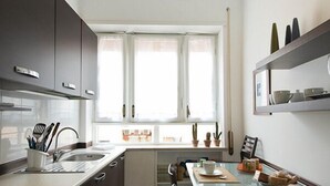 A modern kitchen with dark cabinets, a white countertop, and a yellow ceiling. There is a window with white curtains, a dining table with chairs, and a hanging light fixture. - Minerva B&B (Rome)