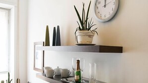 A dining area with a glass table, chairs, and a variety of food items. There is a clock on the wall and a shelf with decorative items and a plant. - Minerva B&B (Rome)