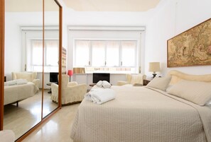 Habitación de hotel con una cama grande, una zona de estar con una silla y una mesita, una televisión y un ventanal con cortinas.