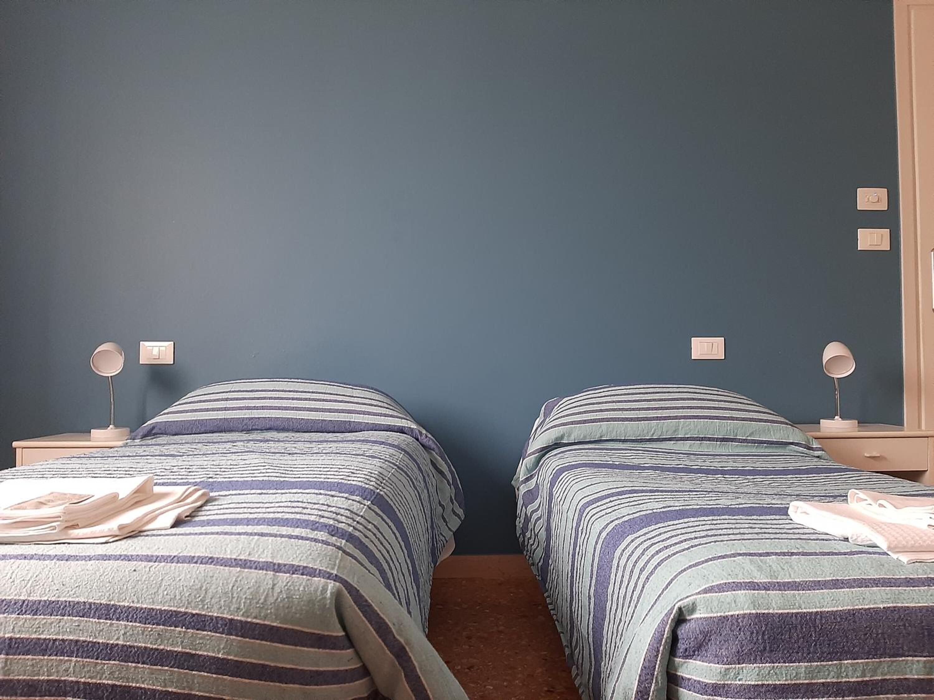 Ein Zimmer mit zwei Betten, jeweils mit gestreifter Bettwäsche und Nachttischlampen. An der dunkelblauen Wand befinden sich zwei fest installierte Schalter.