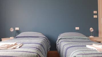 Ein Zimmer mit zwei Betten, jeweils mit gestreifter Bettwäsche und Nachttischlampen. An der dunkelblauen Wand befinden sich zwei fest installierte Schalter.
