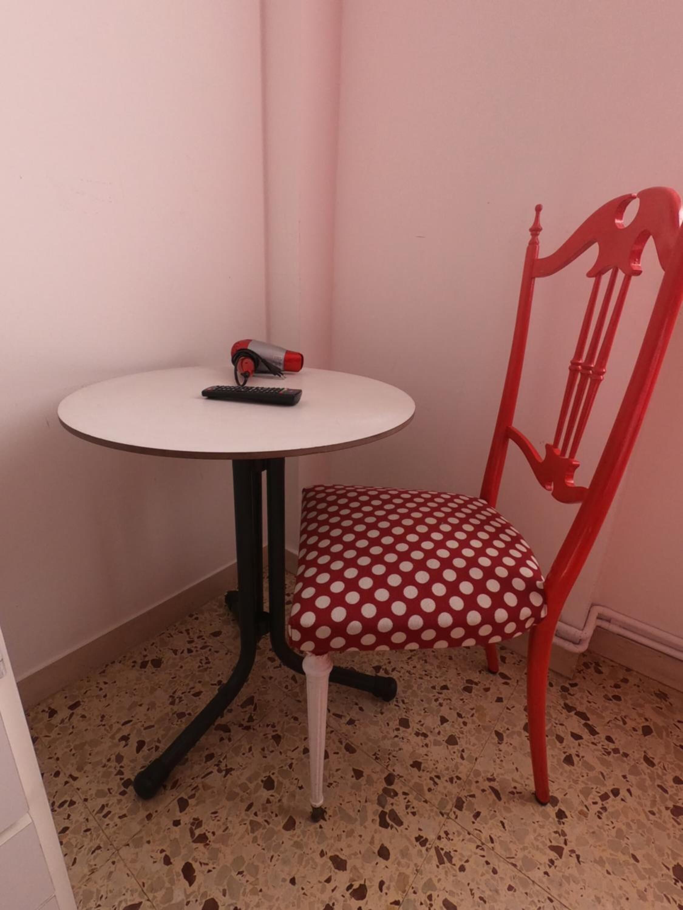 Quarto pequeno com uma mesa branca e uma cadeira vermelha com almofada de bolinhas.