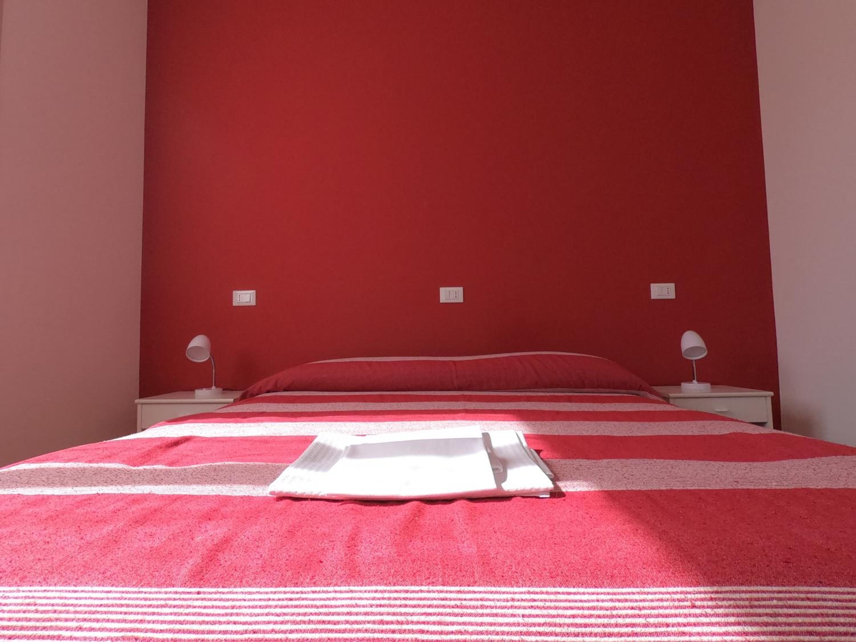 Uma cama com colcha listrada em vermelho e branco, duas luminárias de cabeceira e um criado-mudo branco.