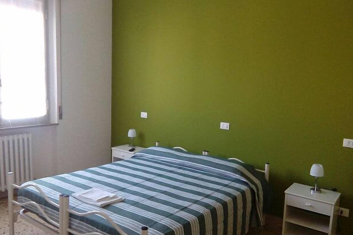 A single-bed room with a green wall, a window, a radiator, a bedside table with a lamp, and a striped bedspread.