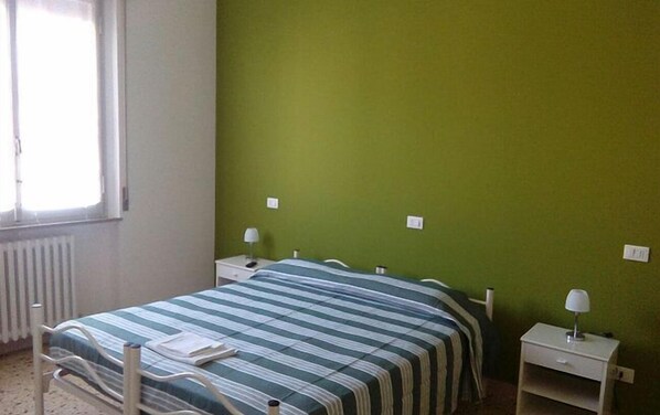 Camera singola con un letto, una parete verde, una finestra, un termosifone, un comodino con una lampada e una coperta a righe.