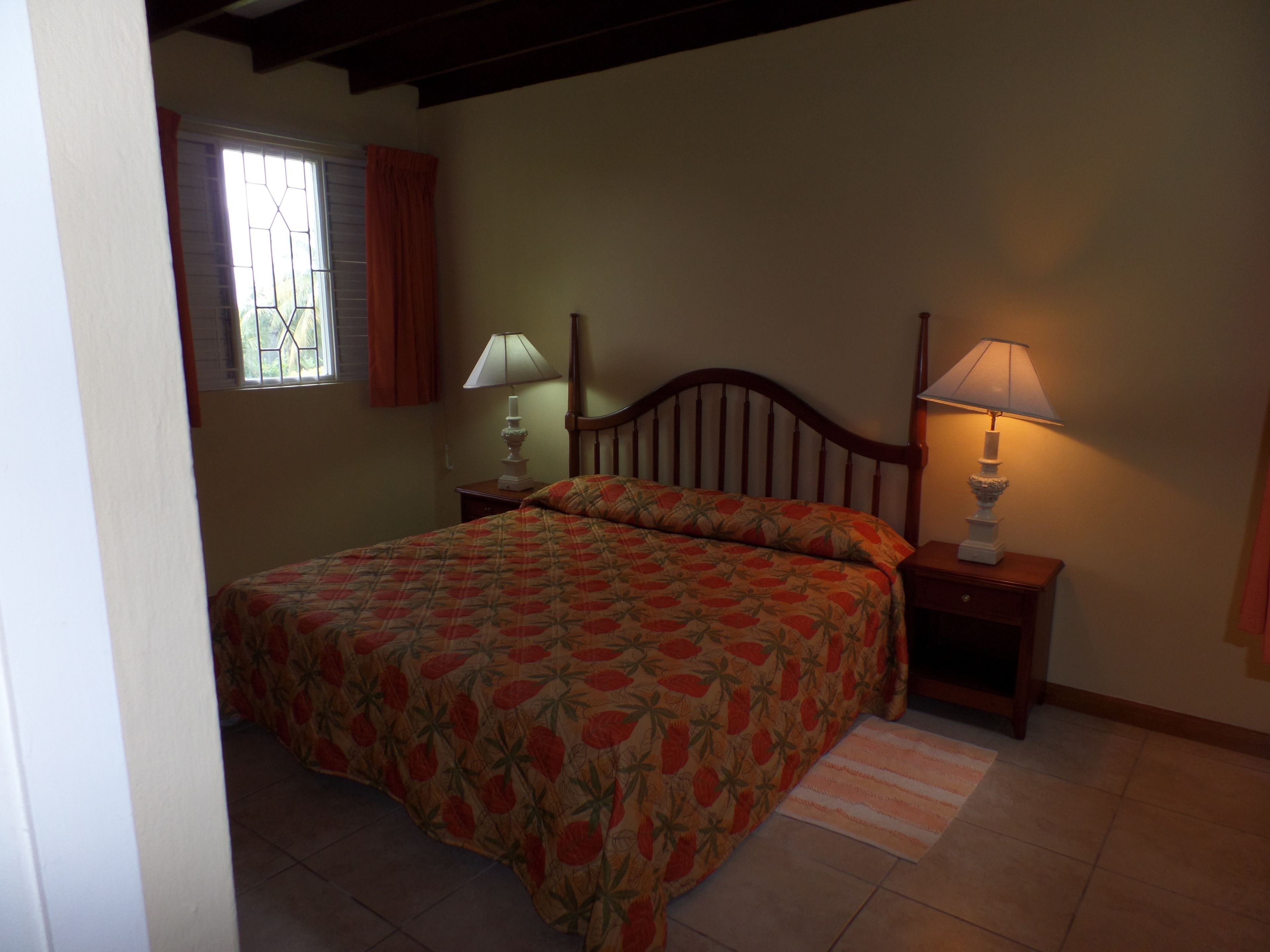 A bedroom with a bed, two bedside tables with lamps, a window with curtains, and a wooden headboard.