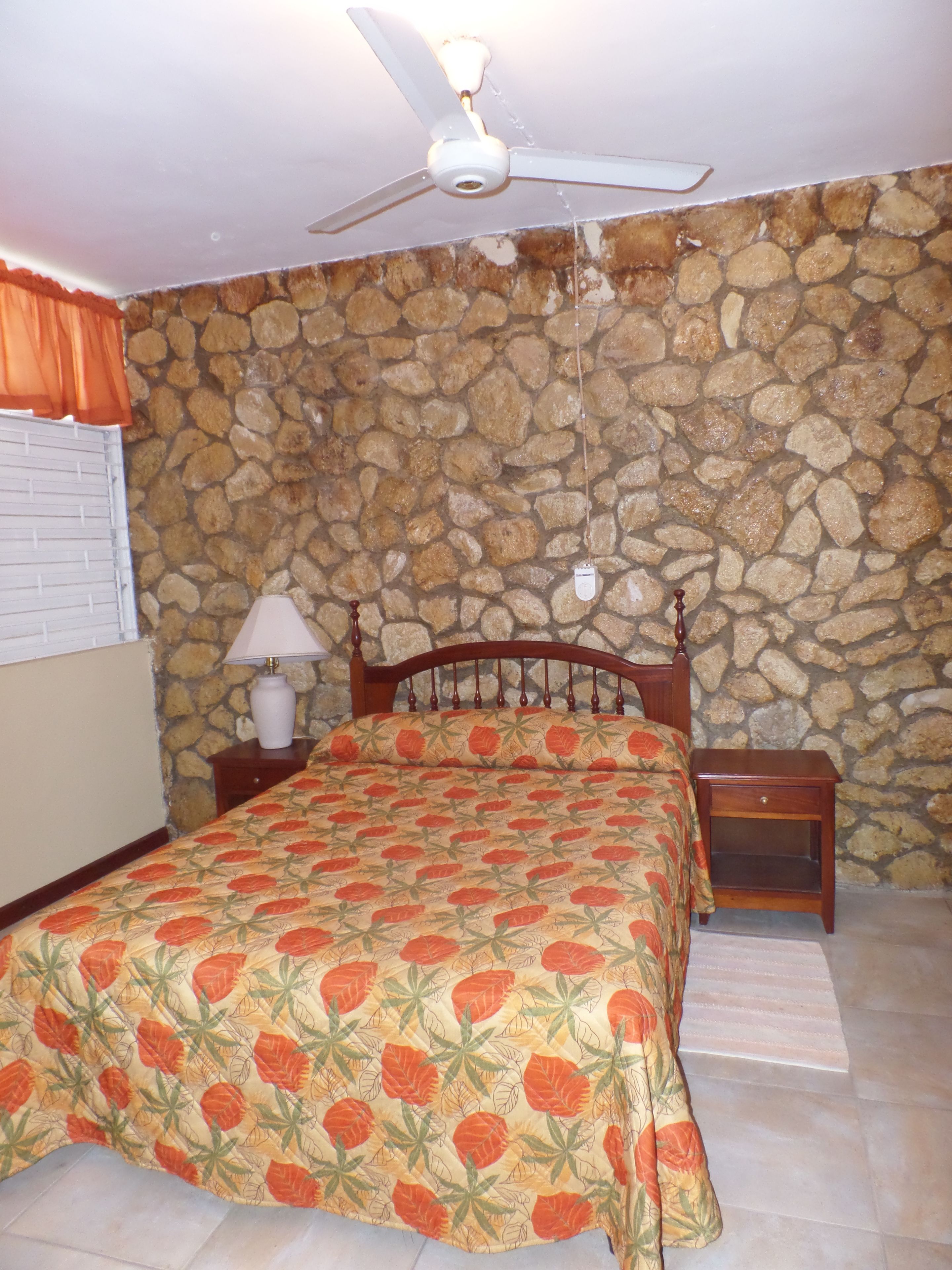 A bedroom with a stone wall, a bed with a floral bedspread, a wooden headboard, a nightstand with a lamp, and a ceiling fan.