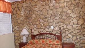 A bedroom with a stone wall, a bed with a floral bedspread, a wooden headboard, a nightstand with a lamp, and a ceiling fan. - Miramar Villa (Ocho Rios)