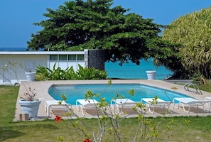 A pool area with white lounge chairs, a stone wall, and a large tree providing shade. - Miramar Villa (Ocho Rios)