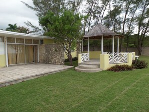 A covered patio area with a stone wall, a small tree, and a gazebo with a white lattice railing. - Miramar Villa (Ocho Rios)