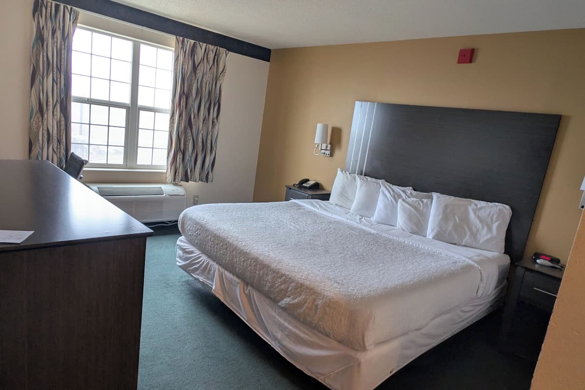 A hotel room with a bed, desk, air conditioning unit, window with curtains, wall-mounted phone, and a nightstand.