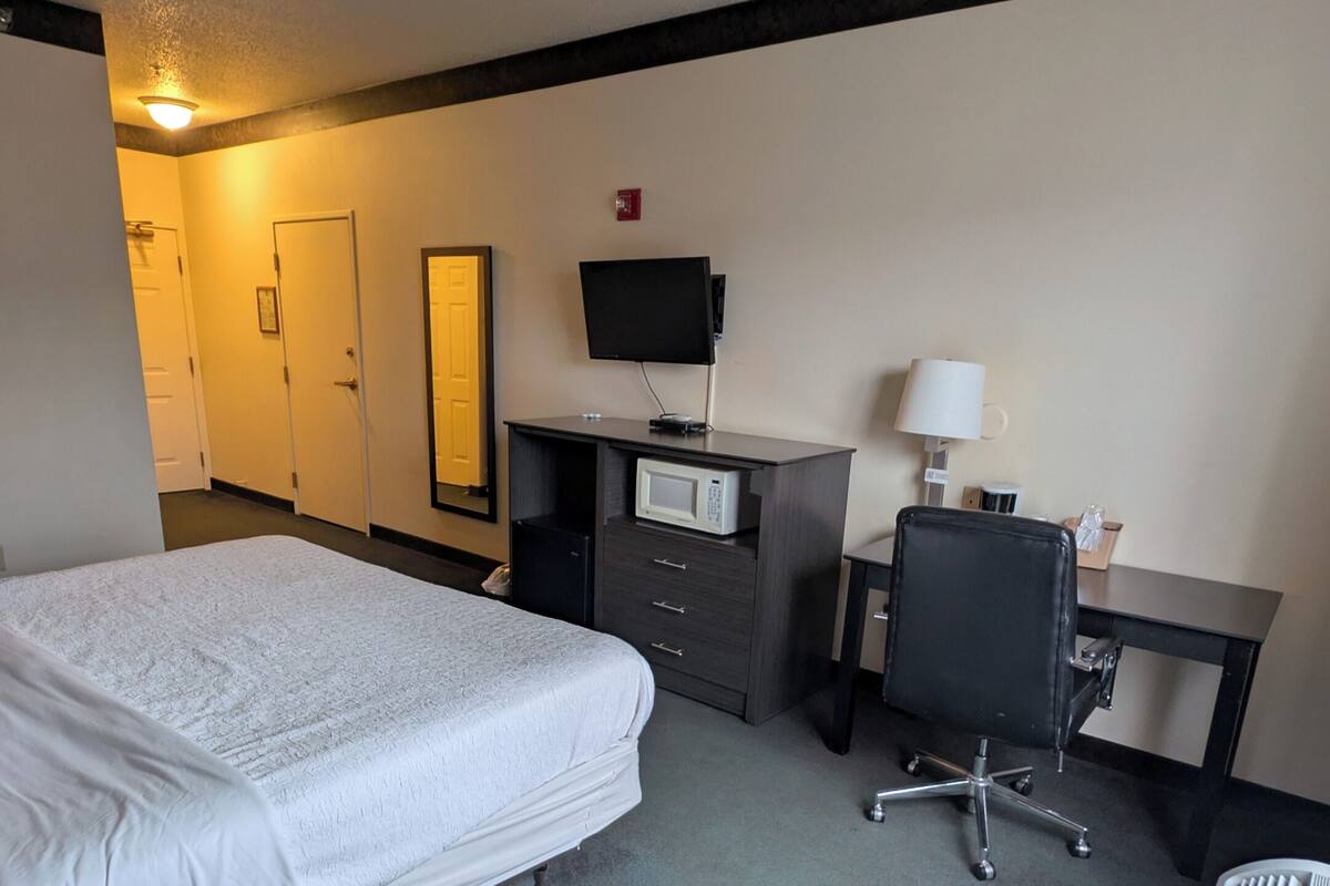 A hotel room with a bed, a desk with a computer, a chair, a microwave, and a heater.