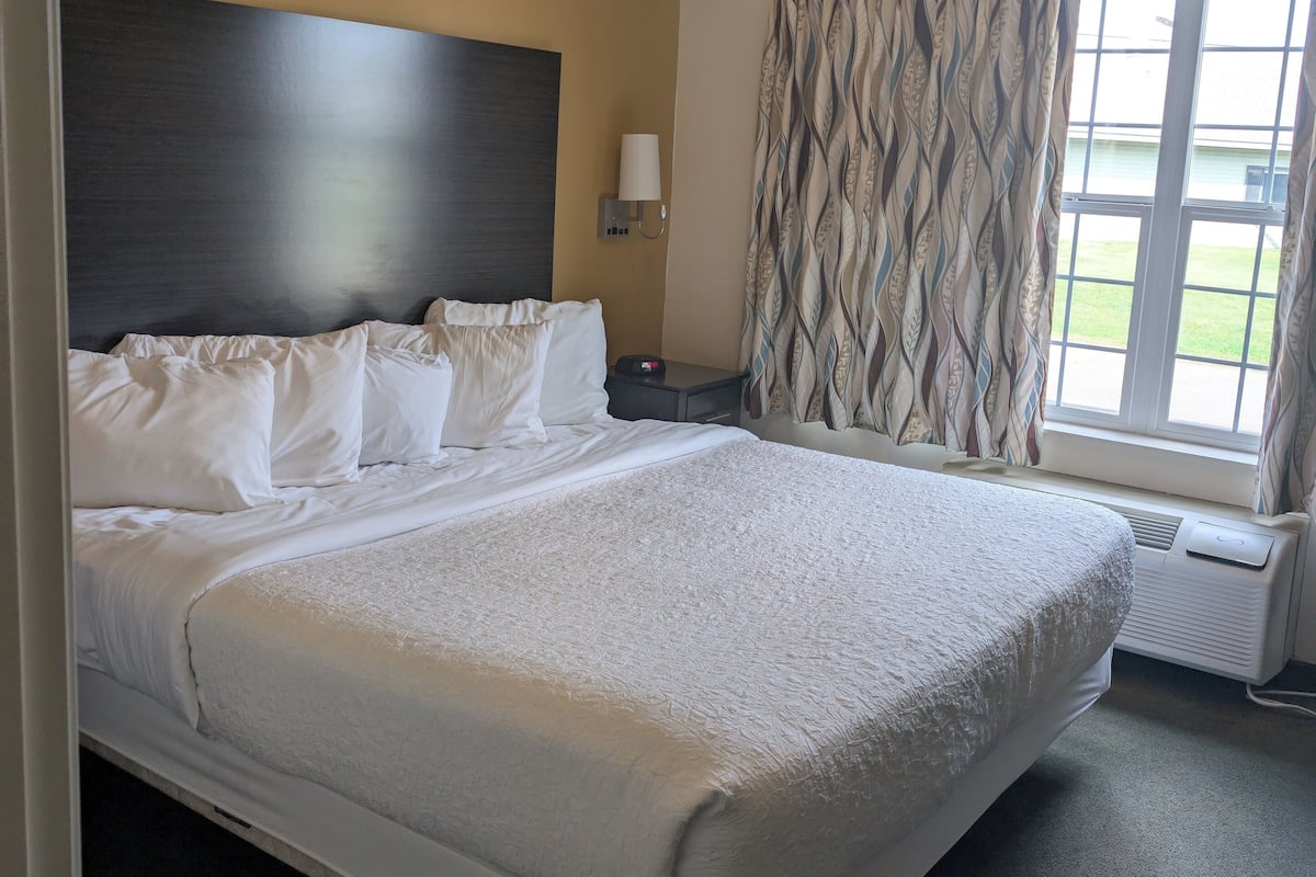 A hotel room with a large bed, a headboard, a bedside table with a lamp, a window with curtains, and a thermostat.