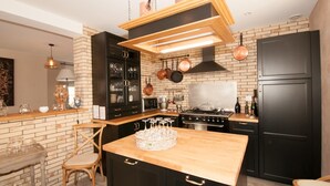 A modern kitchen with black cabinets, a wooden island, hanging pots, and a built-in oven. - Le Logis Aux Bulles (Verzy)