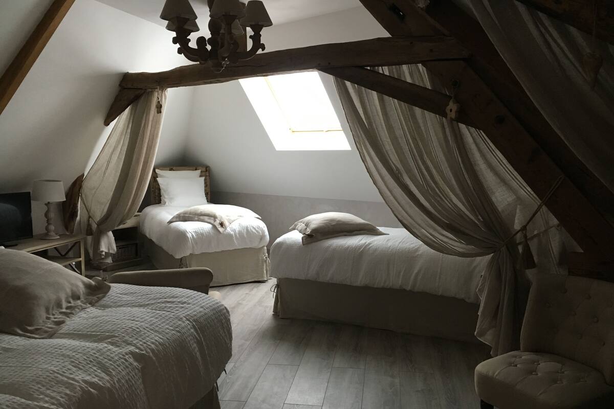 A bedroom with two beds, a wooden ceiling, a window with curtains, and a small table with a lamp.