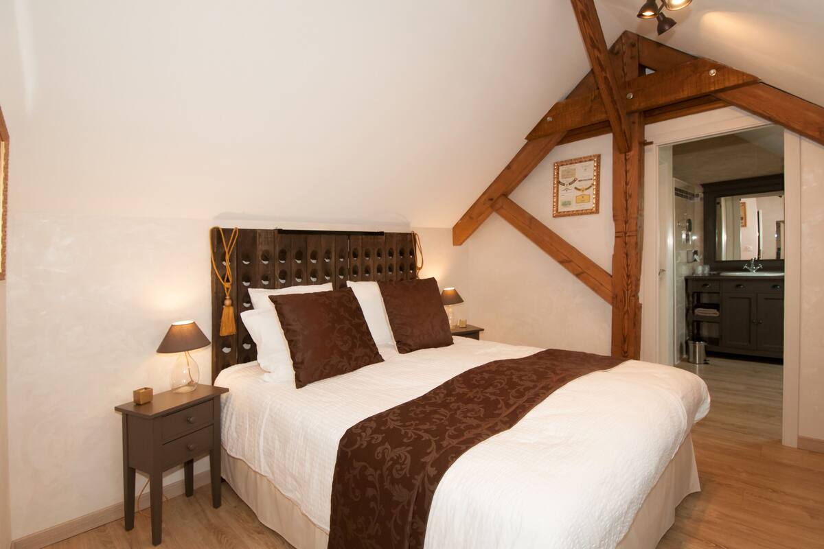 A bedroom with a wooden ceiling, a bed with white and brown bedding, a nightstand with a lamp, and a view into the kitchen.