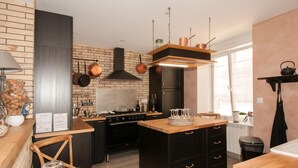 A modern kitchen with a brick wall, black cabinets, copper pots, and a wooden island. - Le Logis Aux Bulles (Verzy)