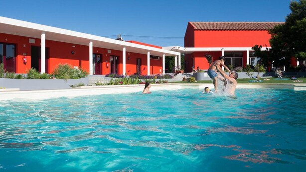 A swimming pool with clear blue water, surrounded by a red building with white columns and a covered area. - Casa D'Edite (Chamusca)