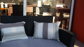 A wicker chair with a cushion and two pillows, positioned near a glass door leading to an interior space with furniture. - Casa D'Edite (Chamusca)