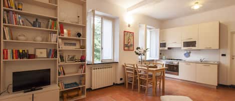 A well-lit, open-plan living and dining area with a kitchen, a bookshelf filled with books, and a television.