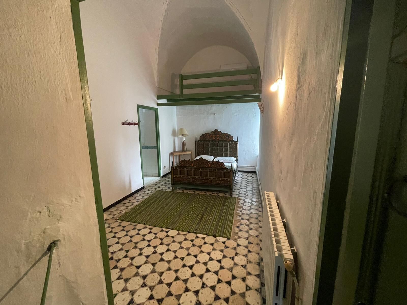 Ein schmaler Flur führt zu einem Schlafzimmer mit einem Bett, einem Nachttisch und einer Lampe. Der Raum hat einen Schachbrettfußboden und eine Heizung.