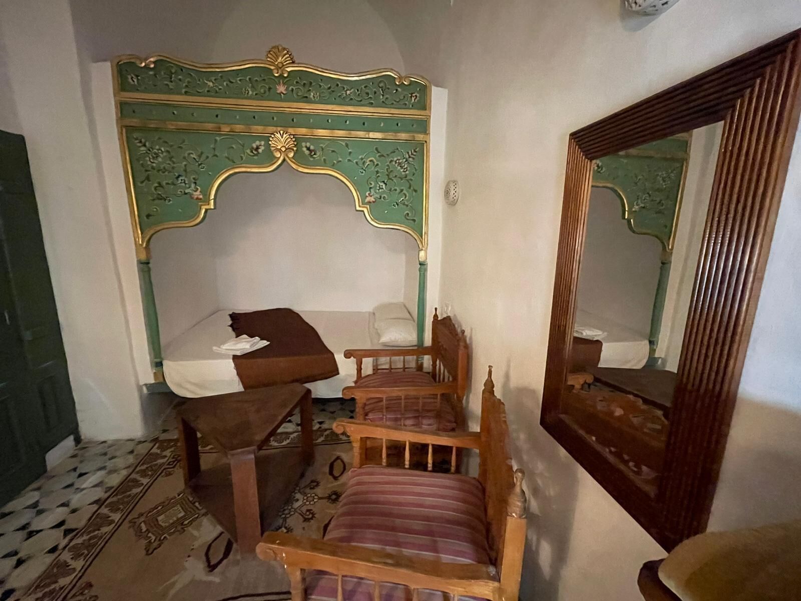 Une chambre à coucher avec une tête de lit verte et dorée, richement décorée, une chaise en bois, un lit recouvert d’une couverture blanche et marron, et un miroir accroché au mur.