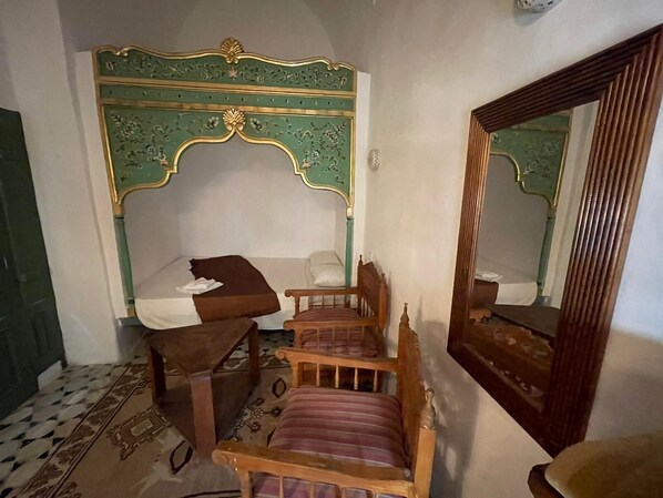 Une chambre à coucher avec une tête de lit verte et dorée, richement décorée, une chaise en bois, un lit recouvert d’une couverture blanche et marron, et un miroir accroché au mur.