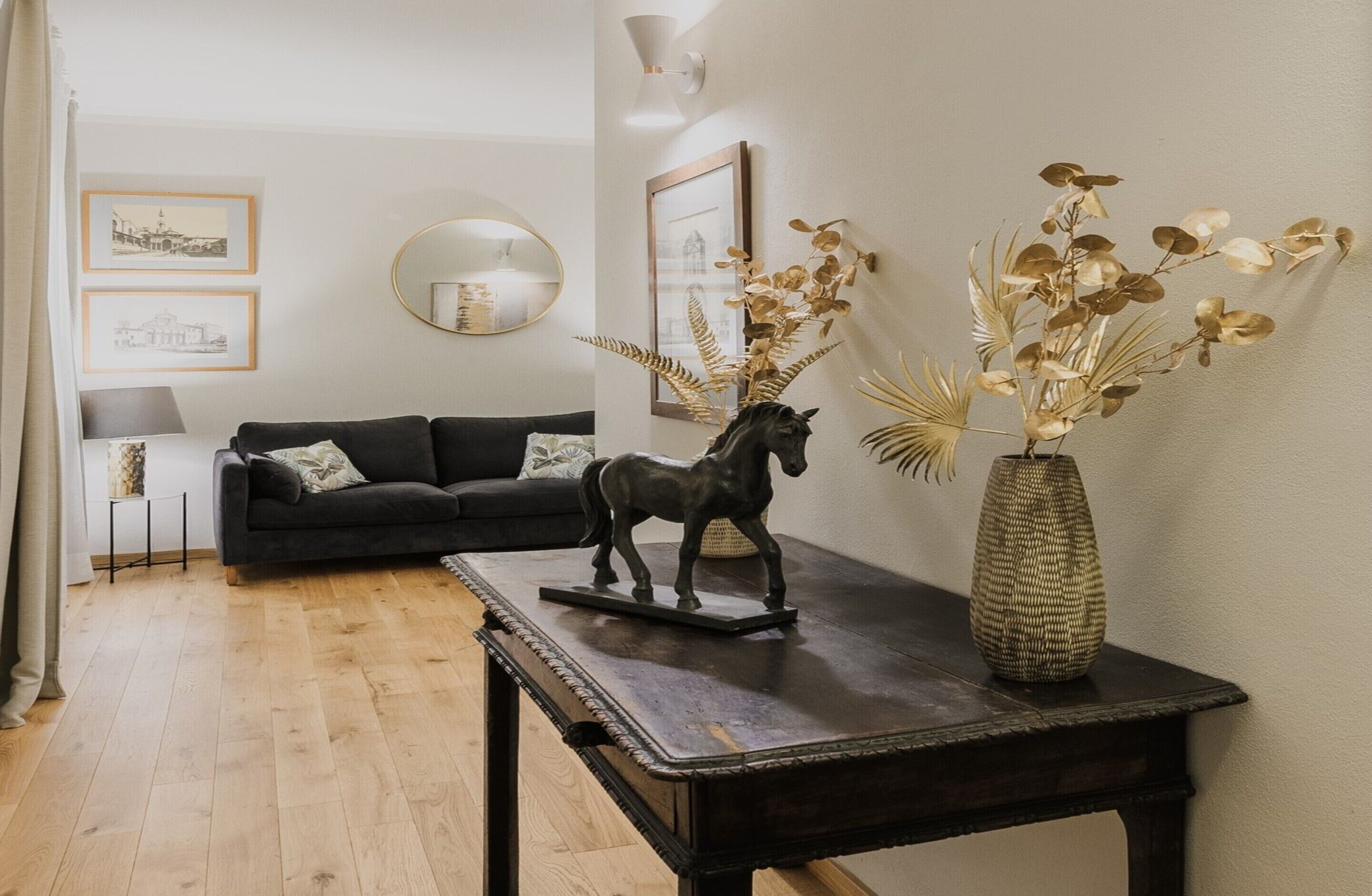 A living room with a dark sofa, a small table with a horse figurine and a vase of dried plants, and framed pictures on the wall.