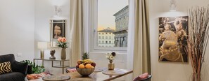 A modern living room with a dining area, featuring a large window with a city view, a fruit bowl on the table, and a painting on the wall. - Appartamenti La Medicea (Florence)