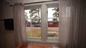 Une pièce avec une table et des chaises en bois, une fenêtre avec des rideaux et une vue sur un bâtiment rouge et une voiture à l’extérieur.