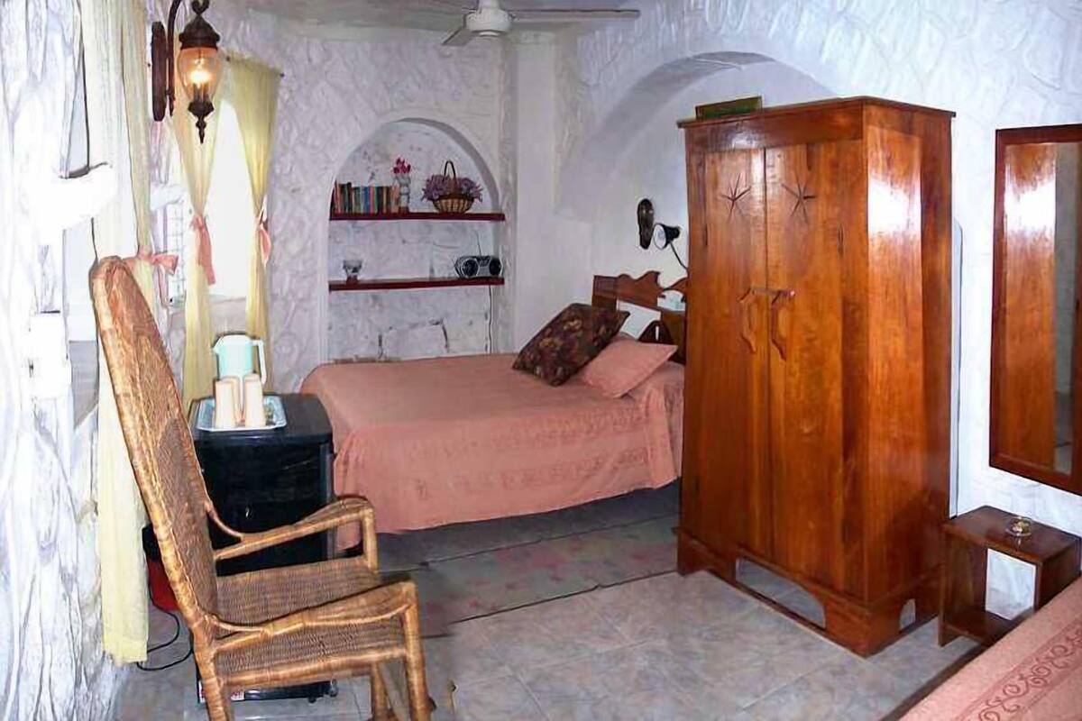 Une chambre à coucher comprenant un lit, une armoire en bois, une chaise en osier, une petite table et une étagère avec des livres et des objets décoratifs.