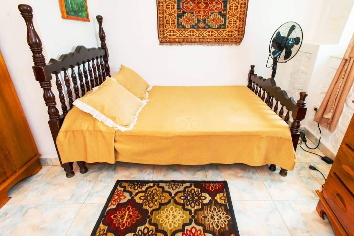 Une chambre avec un lit en bois, un tapis à motifs, une décoration murale et un ventilateur.