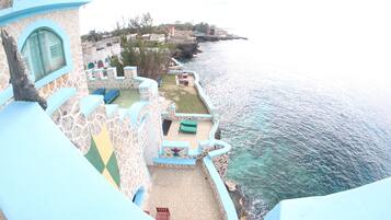 A seaside hotel with a terrace overlooking the ocean, featuring a pool, lounge chairs, and a dining area.