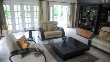 A living room with a sofa, armchair, and coffee table, a kitchen area with appliances, and large windows with curtains.
