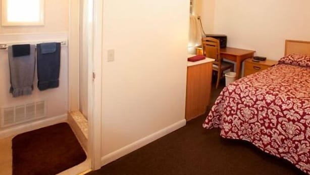A small, well-lit bedroom with a bed, a desk, and a chair. There is a door with a towel hanging on it, and a small rug on the floor. - Charlie's Motel (Frostburg)
