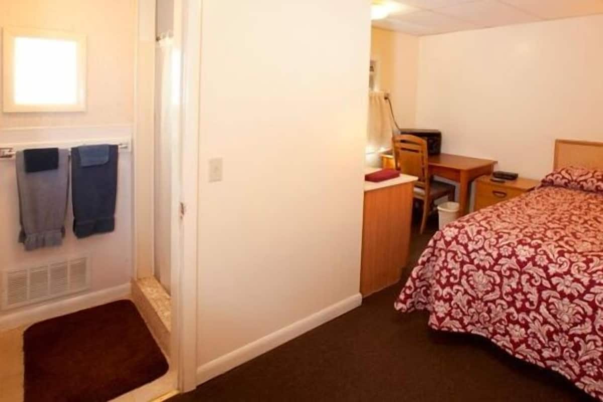 A small, well-lit bedroom with a bed, a desk, and a chair. There is a door with a towel hanging on it, and a small rug on the floor.