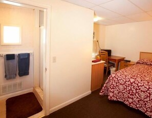 A small, well-lit bedroom with a bed, a desk, and a chair. There is a door with a towel hanging on it, and a small rug on the floor. - Charlie's Motel (Frostburg)