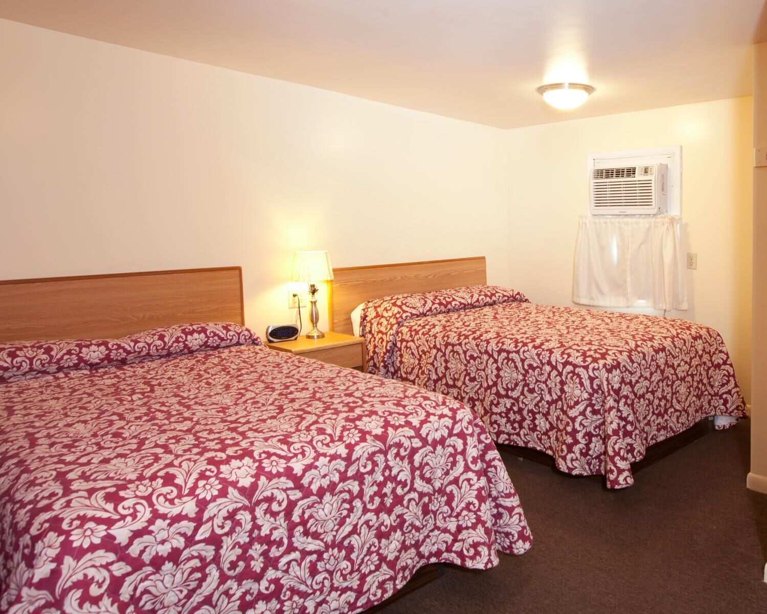 A hotel room with two beds, a nightstand with a lamp, an air conditioning unit, and a window with curtains.