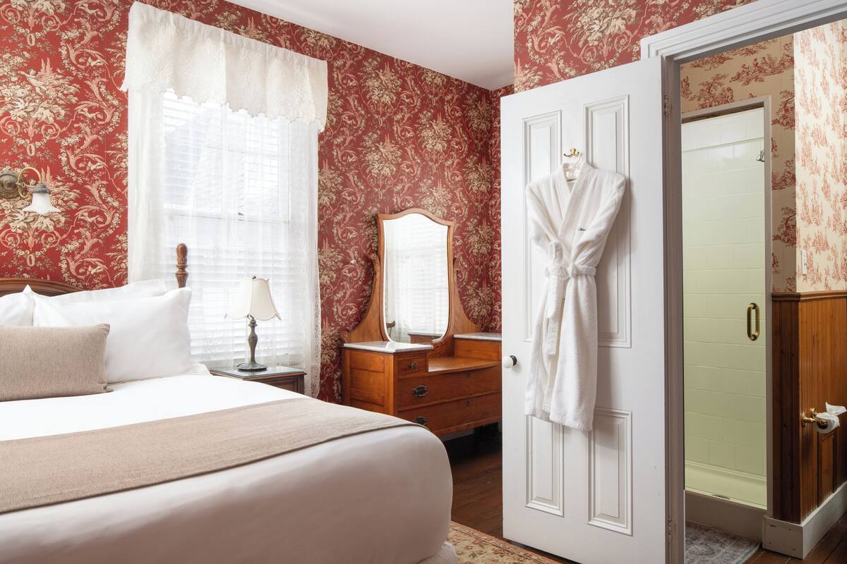 A hotel room with a bed, a wooden dresser with a mirror, a robe hanging on the door, and a shower area.