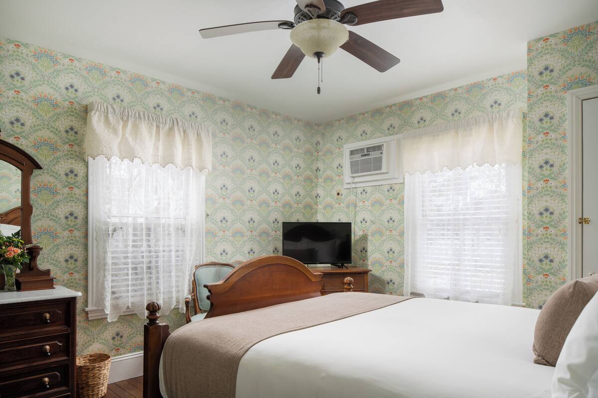 A bedroom with floral wallpaper, a bed, a wooden dresser, a television, and a ceiling fan.