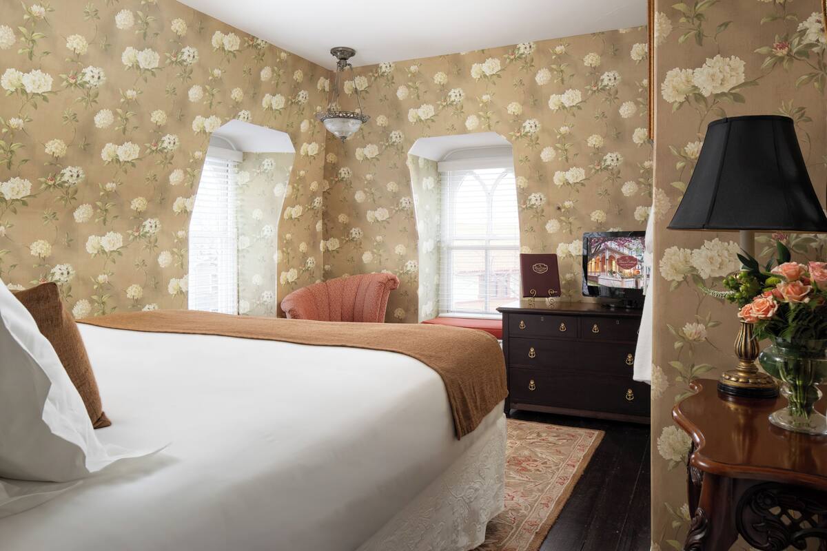 A bedroom with floral wallpaper, a bed with white linens, a dresser, and a lamp.