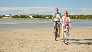 Two people riding bicycles on a sandy beach with a body of water and buildings in the background.