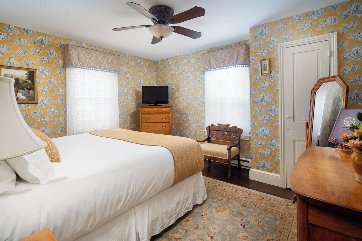 A bedroom with a bed, dresser, television, chair, and a ceiling fan.