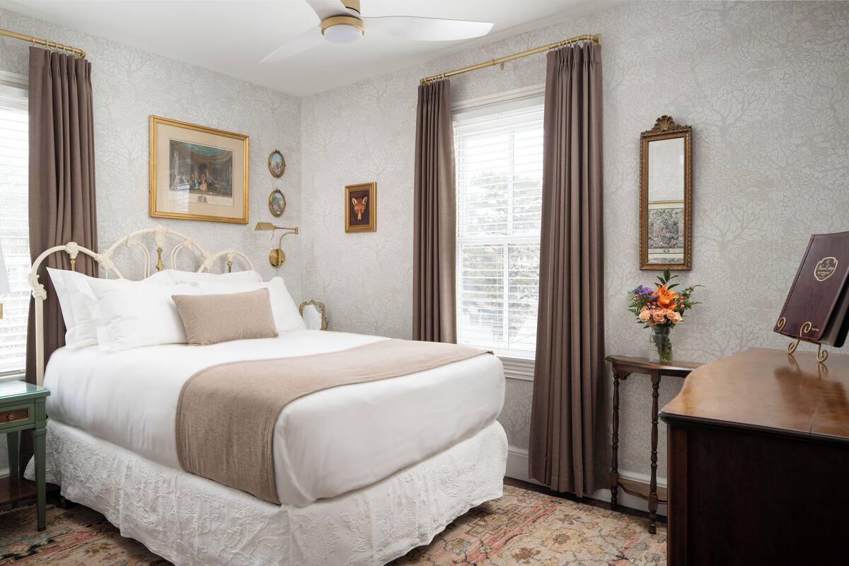 A neatly arranged bedroom with a large bed, a bedside table, a mirror, and a window with curtains.