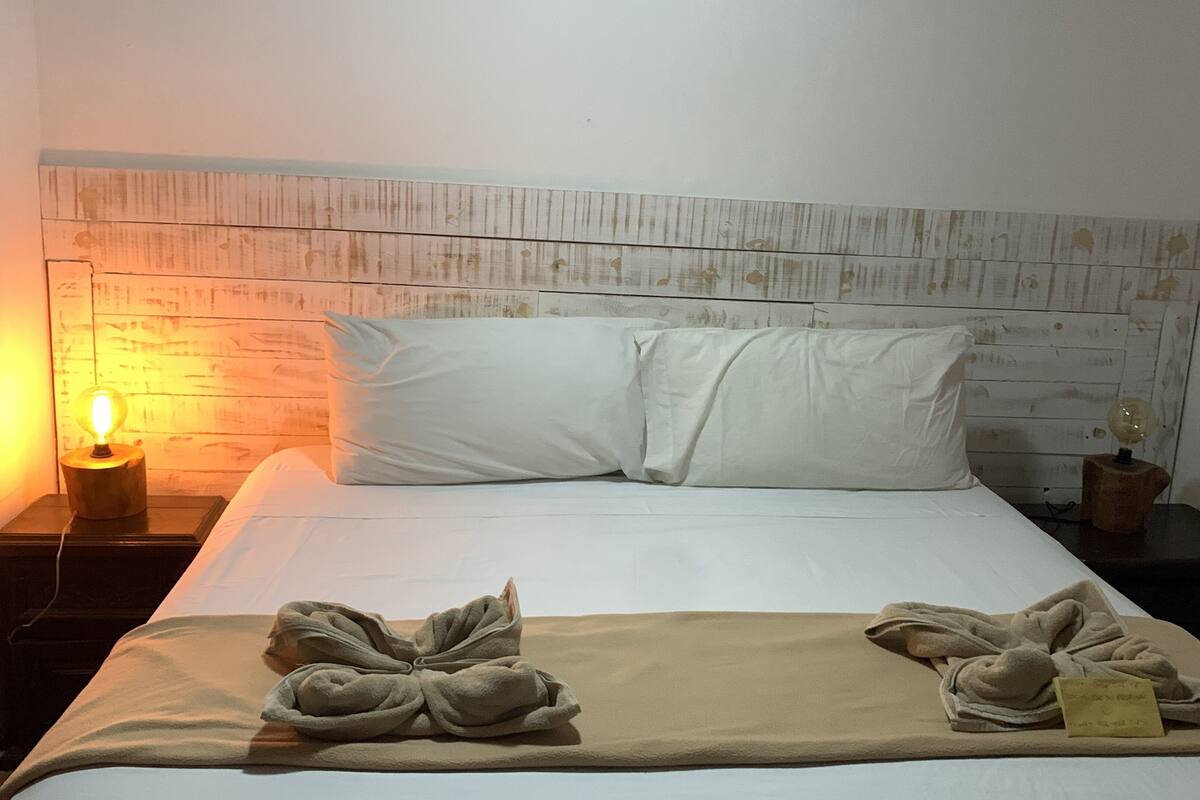 Ein ordentlich gemachtes Bett mit weißen Leinentüchern, zwei Kissen und einer beige farbenen Decke, die am Fuß des Bettes gefaltet ist. Es gibt zwei Nachttische, jeder mit einer Lampe und einem kleinen dekorativen Gegenstand. Das Kopfteil hat eine rustikale Holzoptik.