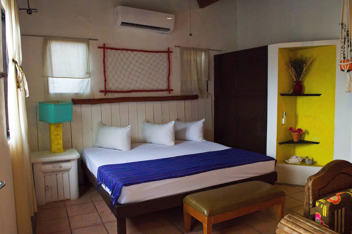 Ein Schlafzimmer mit Bett, einer Bank, einem Nachttisch, einer Lampe, einem Fenster und einem Wandregal mit Dekoration.