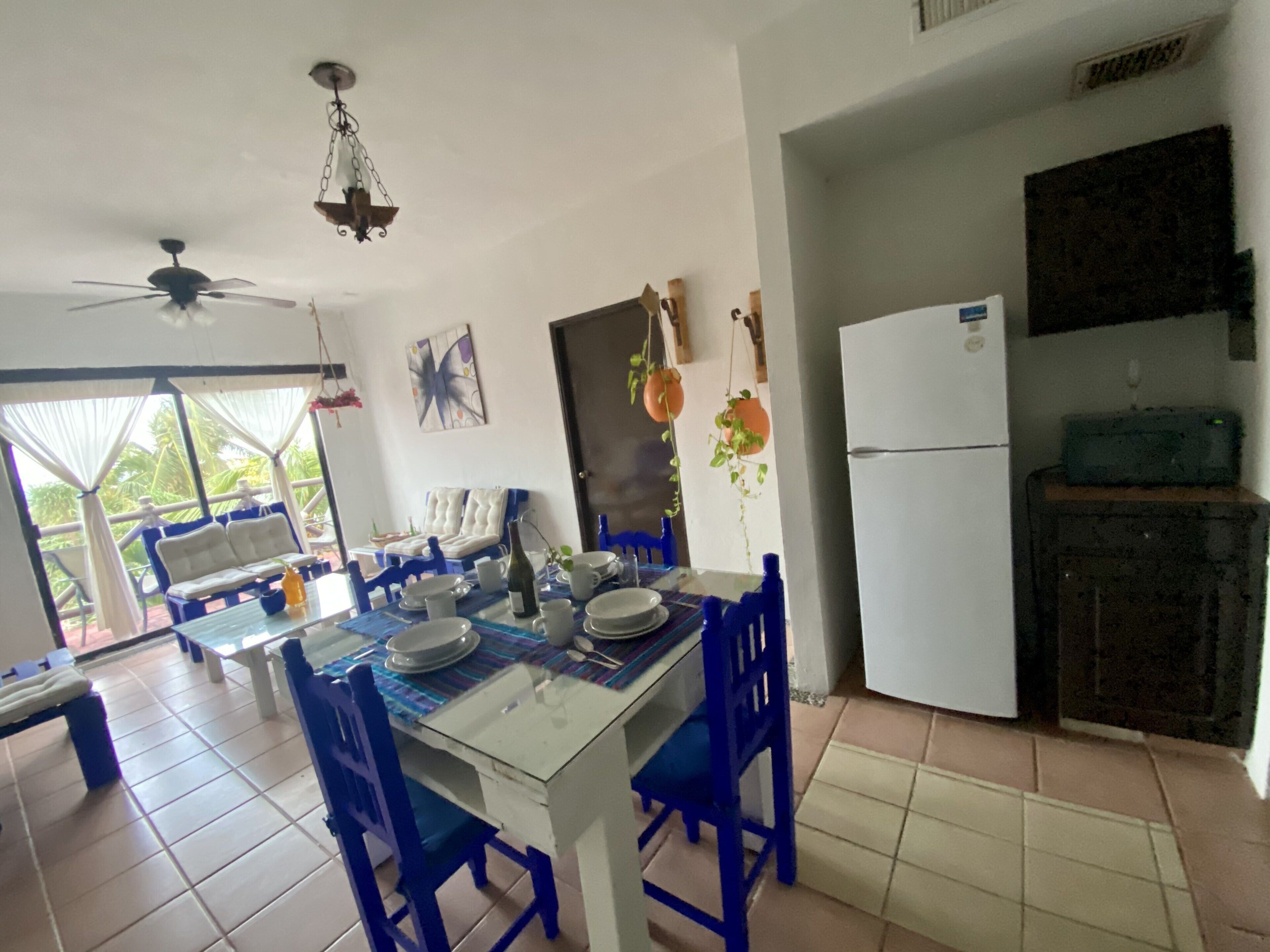 A dining area with a table set for four, blue chairs, a ceiling fan, and a refrigerator.