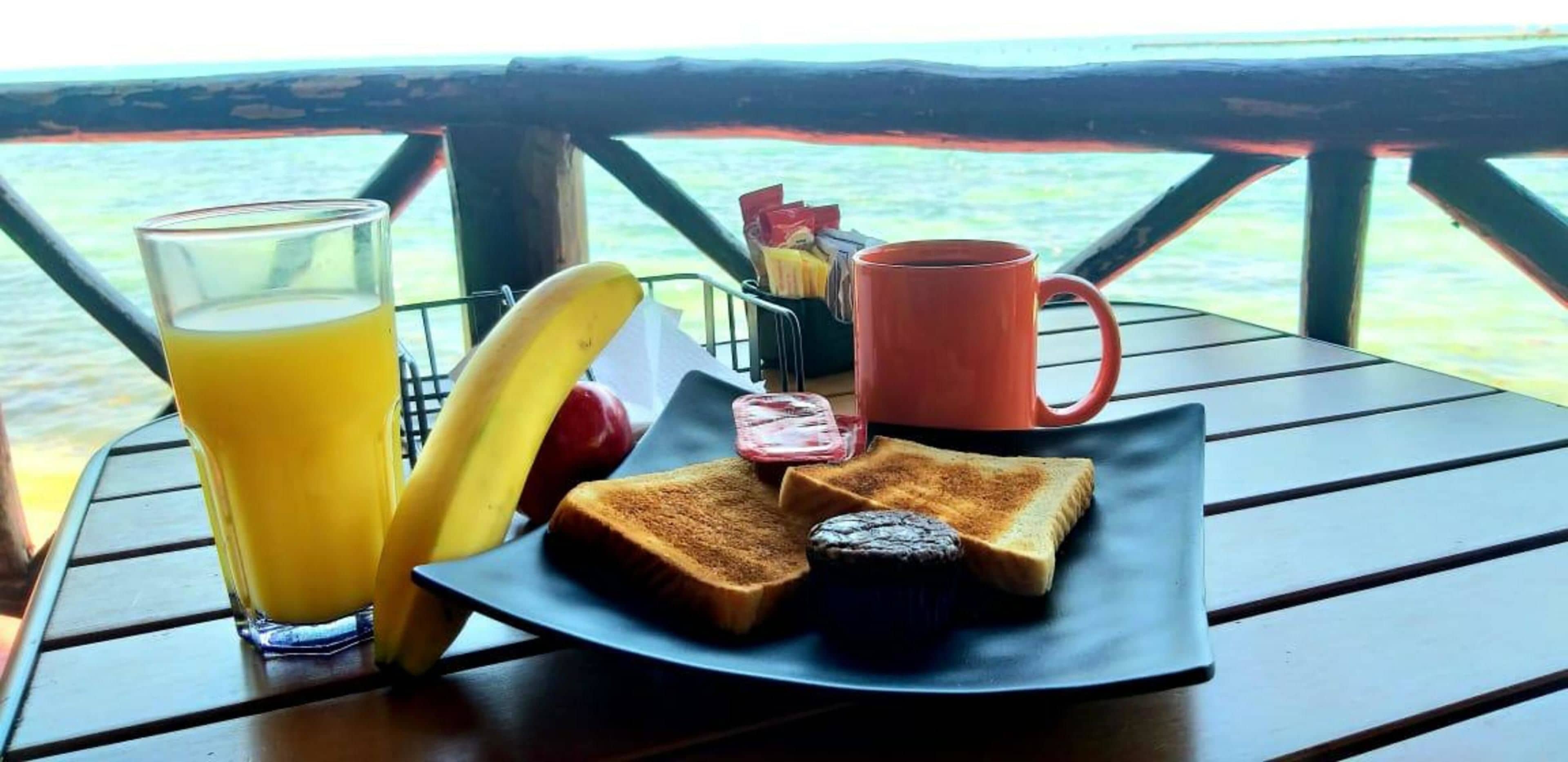 Ein Frühstücksbrett mit Toast, Marmelade, einem Muffin, einem Glas Orangensaft und einer roten Tasse auf einem Holztisch mit Blick aufs Meer.