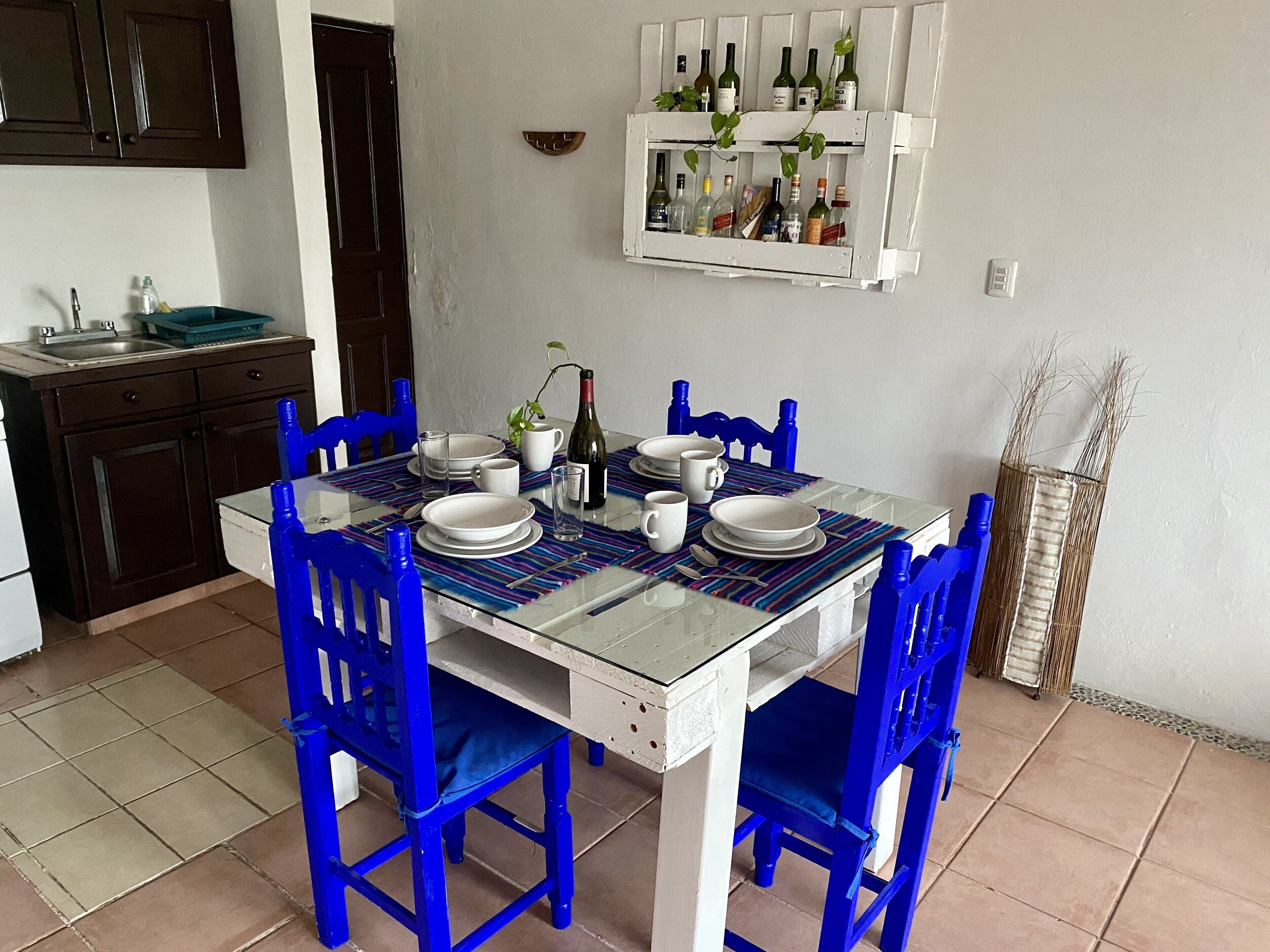 Een eethoek met een glazen tafel, blauwe stoelen, witte borden en een wijnrek aan de muur.