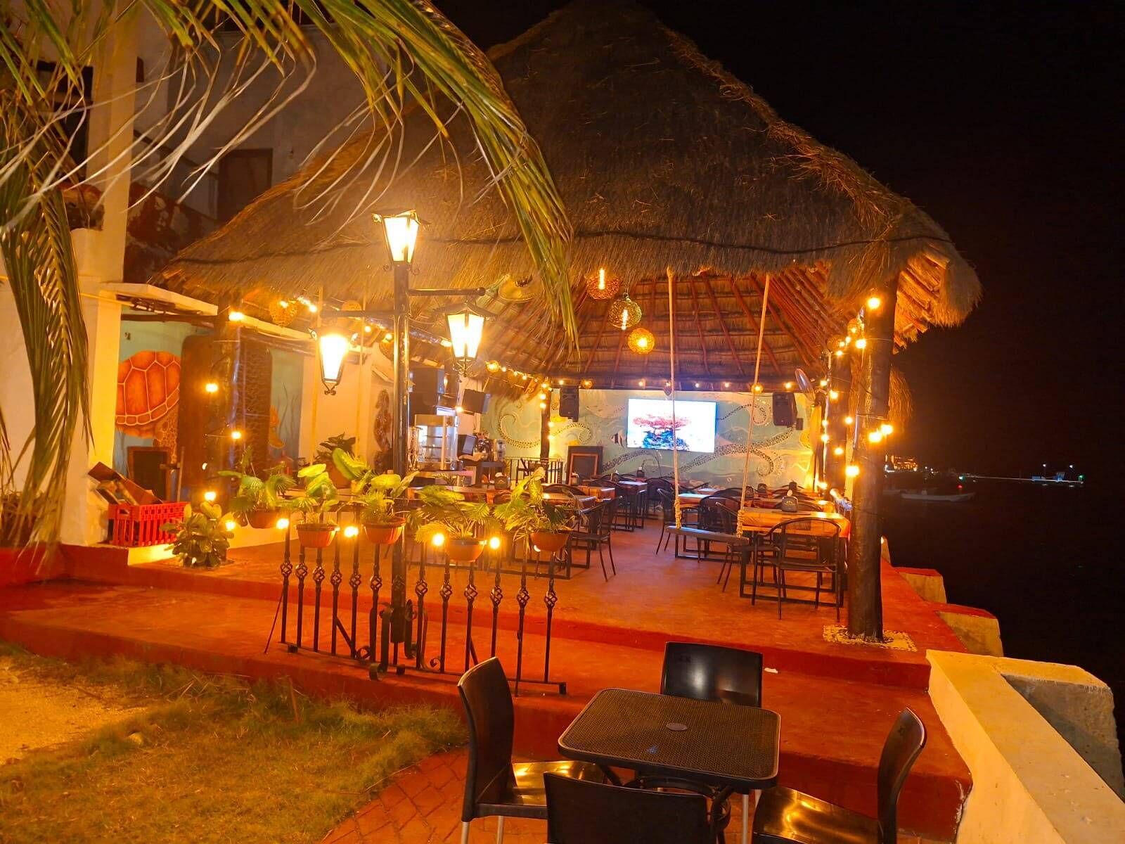 Un área de comedor al aire libre con mesas y sillas de techo de paja, rodeada de plantas en macetas y luces colgantes.